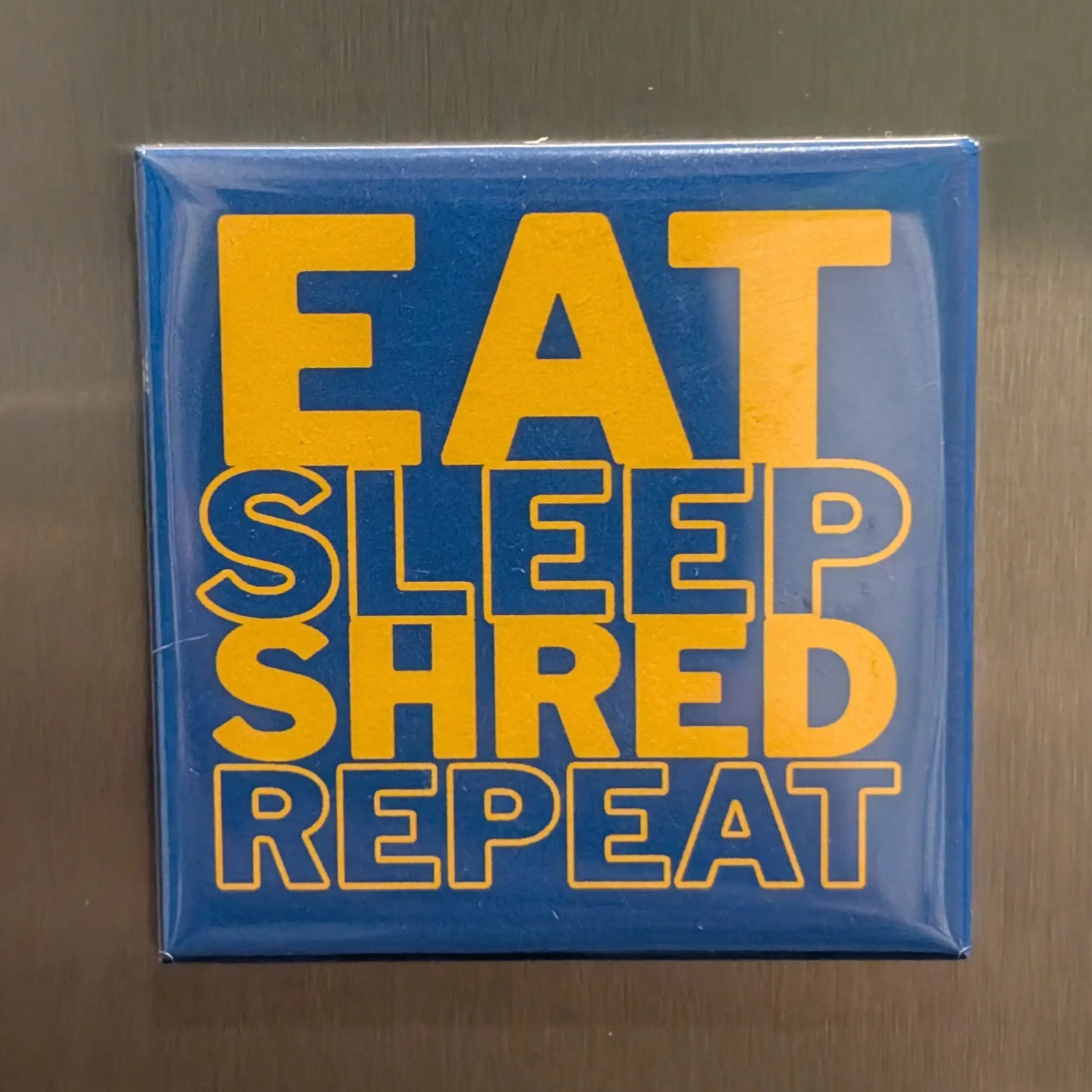 Blue Fridge Magnet with yellow text 'EAT SLEEP SHRED REPEAT' on a reflective surface