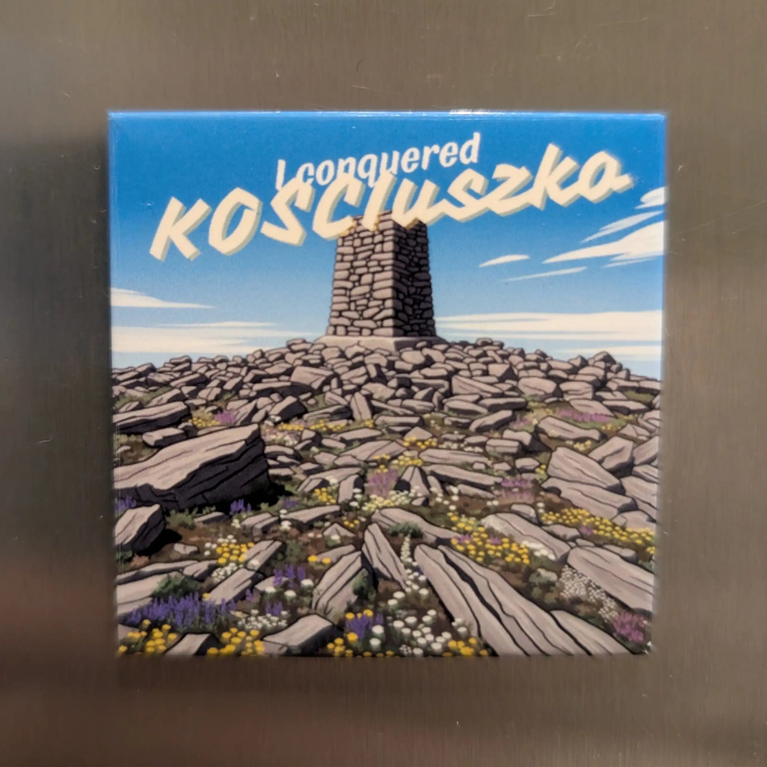 Fridge magnet with a mountain summit design and text on a metallic surface