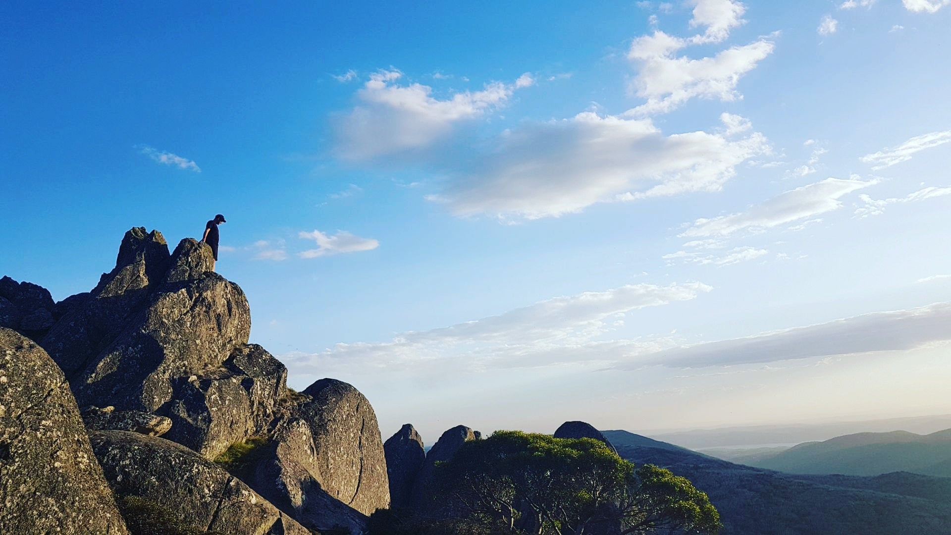 Person standing on a rocky mountain peak with a clear blue sky and distant mountains.