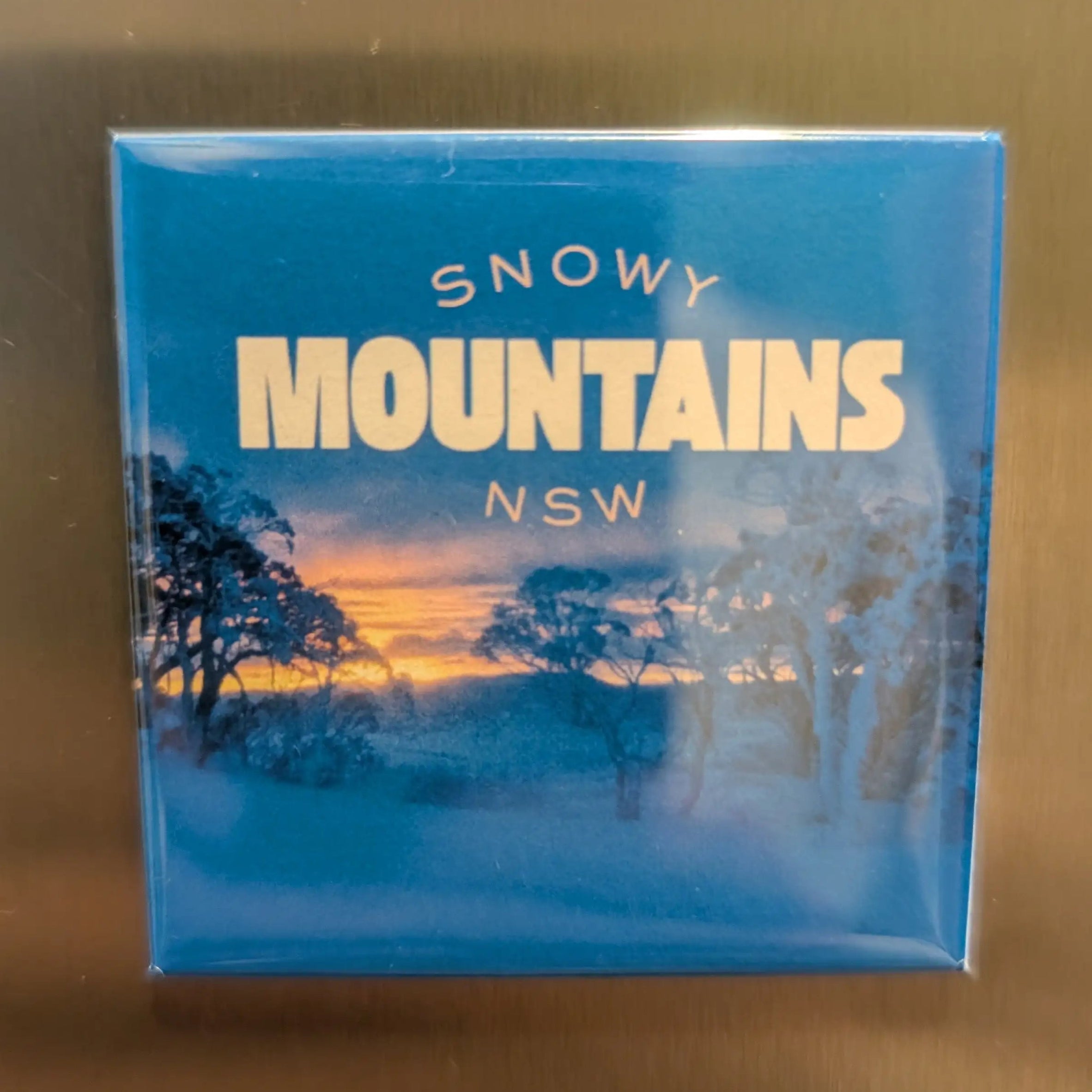 Rectangular magnet with 'Snowy Mountains NSW' text and scenic image on a reflective surface.